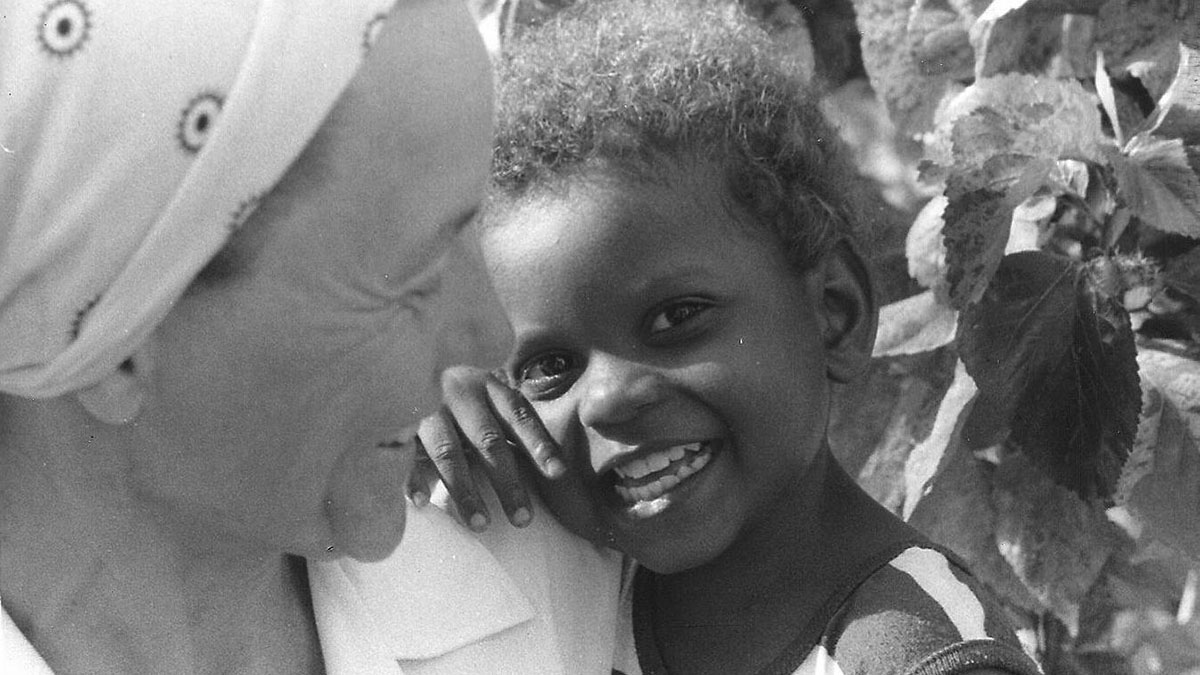 Black-and-white photo of a smiling woman in a headscarf embracing a joyful child in Jonestown, Guyana.