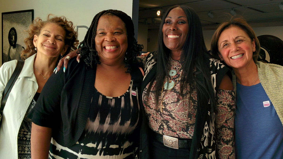 Four women smiling and standing arm-in-arm at the Museum of the African Diaspora during a 2017 panel on Jonestown remembrance.